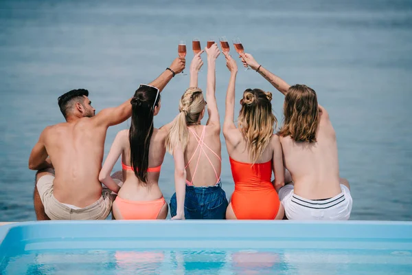Vista posteriore di giovani amici di sesso maschile e femminile bicchieri di champagne mentre seduto vicino alla piscina in spiaggia — Foto stock
