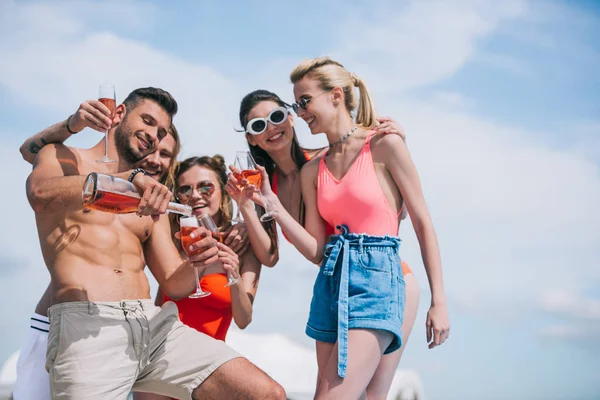 Low angle view of happy young friends holding glasses and pouring wine at summer day — Stock Photo