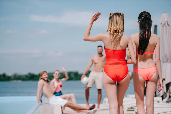 Вид ззаду на дівчат у купальнику, що ходить на пляжі і махає рукою друзям на березі басейну — стокове фото