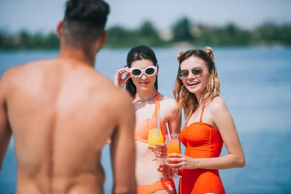 Selective focus of girls in swimwear holding cocktails and looking at young man on beach — Stock Photo