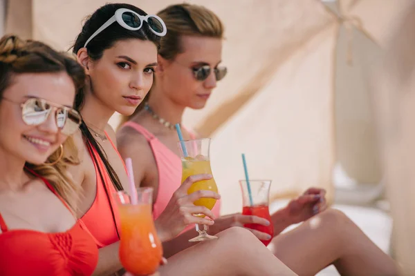 Atractivas novias en traje de baño y gafas de sol bebiendo cócteles en bungalow en la playa - foto de stock