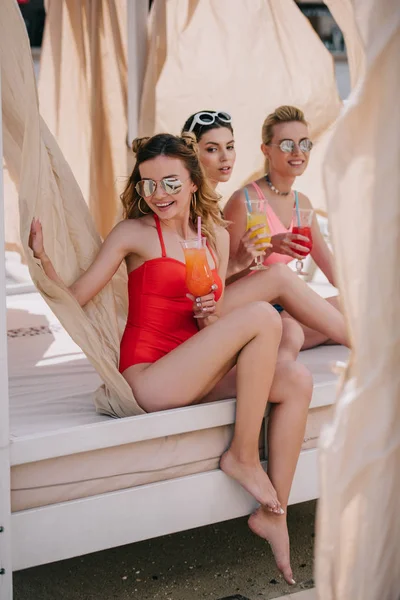 Amigas felices en traje de baño y gafas de sol beber cócteles en bungalow en la playa - foto de stock