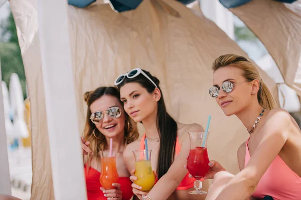 Schöne junge Frauen in Badebekleidung und Sonnenbrille trinken Cocktails in Bungalow am Strand — Stockfoto