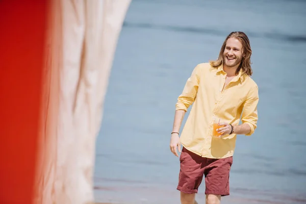 Giovane uomo sorridente che tiene il bicchiere con un cocktail e distoglie lo sguardo mentre cammina sulla spiaggia — Foto stock