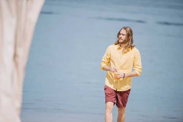 Schöner junger Mann hält Glas mit Cocktail und schaut weg, während er am Strand spaziert — Stockfoto