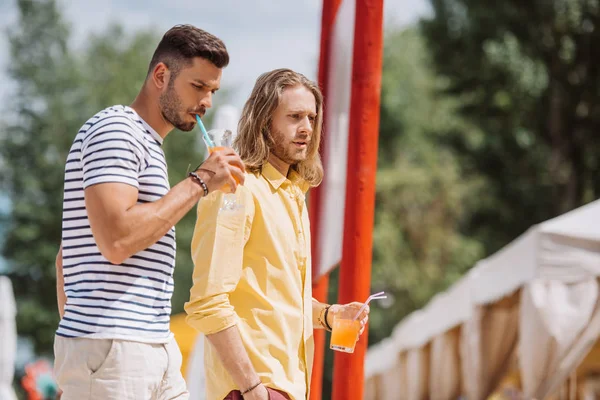 Gutaussehende junge Männer, die Gläser mit Cocktails halten und gemeinsam am Strand spazieren gehen — Stockfoto