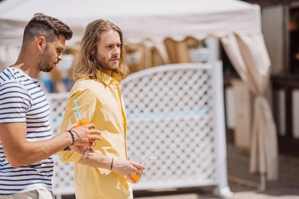 Vue latérale de beaux jeunes hommes tenant des lunettes avec des cocktails et marchant ensemble sur la plage — Photo de stock