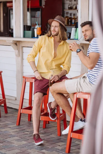 Молодые друзья-мужчины пьют пиво и смотрят в сторону, сидя вместе в пляжном баре — стоковое фото