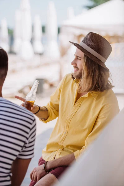 Jeune homme tenant une bouteille de bière et pointant du doigt tout en étant assis avec un ami au bar de la plage — Photo de stock