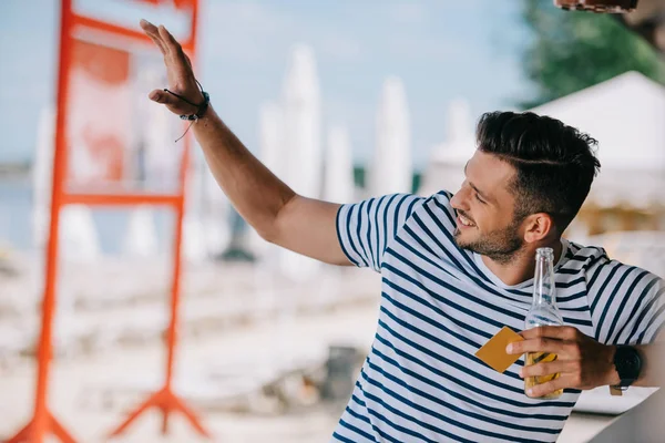 Handsome smiling young man holding beer bottle and credit card while waving hand and looking away — Stock Photo