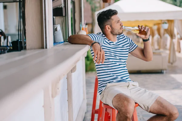 Молодой человек пьет пиво из стеклянной бутылки, сидя в баре на пляже — стоковое фото