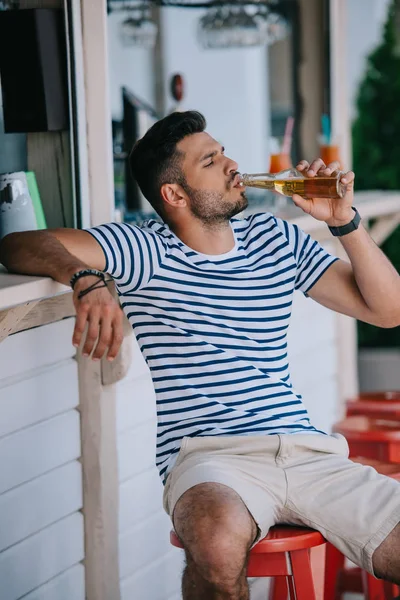 Joven guapo bebiendo cerveza de botella de vidrio mientras está sentado en el bar de la playa — Stock Photo