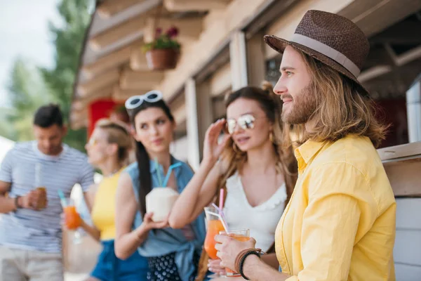 Vue latérale du jeune homme souriant regardant ailleurs tout en buvant des cocktails avec des amis au bar de la plage — Photo de stock