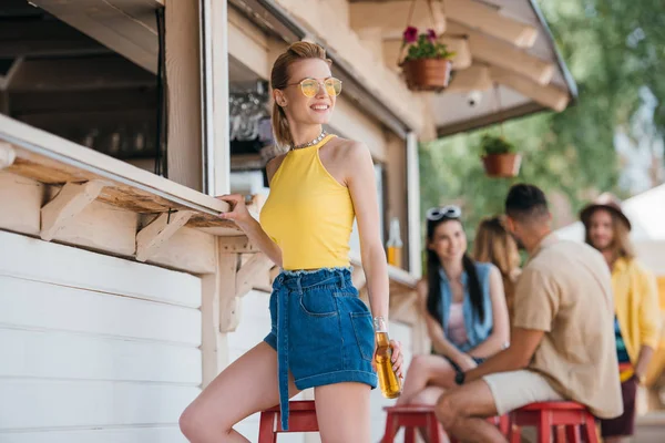 Smiling young woman holding beer bottle while friends sitting behind at beach bar — Stock Photo