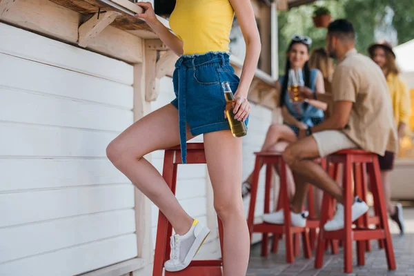 Recortado disparo de chica sosteniendo botella de cerveza mientras amigos sentados detrás en el bar de la playa - foto de stock