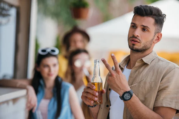 Jeune homme tenant une bouteille de bière, geste avec les doigts et regardant loin au bar de la plage — Photo de stock