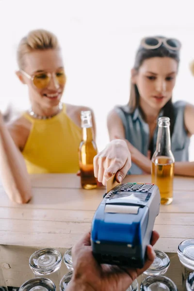 Selective focus of girls drinking beer and paying with credit card on terminal in beach bar — Stock Photo