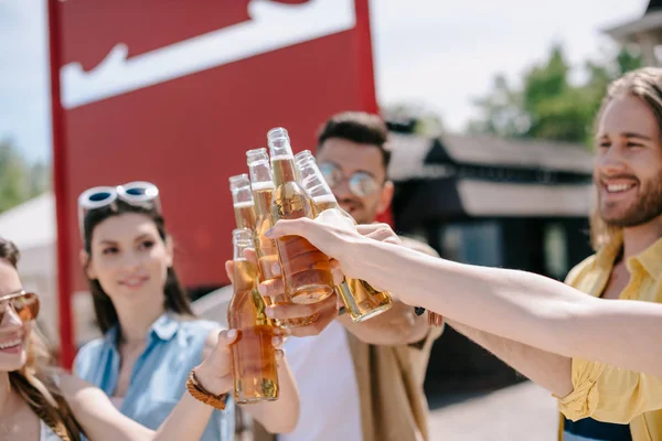 Recortado disparo de jóvenes sonrientes tintineo botellas de cerveza en el día de verano - foto de stock