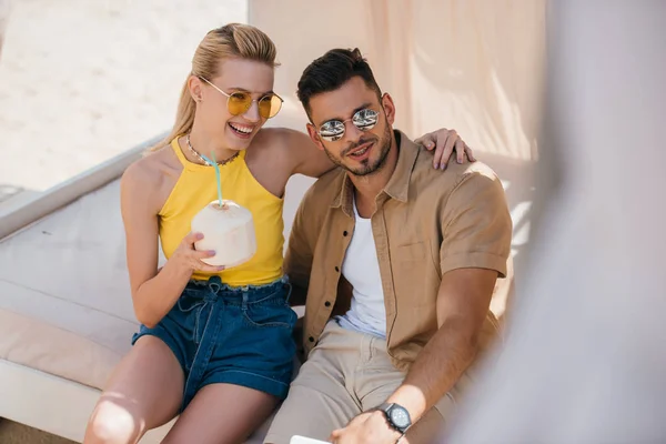 Счастливая молодая пара в солнечных очках, пьющая летний коктейль, сидя вместе в бунгало на пляже — стоковое фото