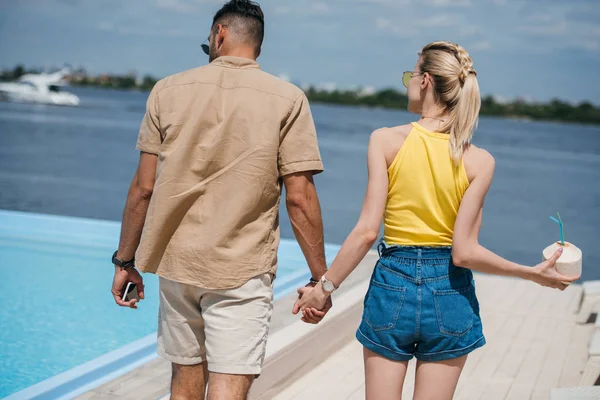 Vista trasera de pareja joven con cóctel de verano y teléfono inteligente cogido de la mano y caminando en la playa - foto de stock