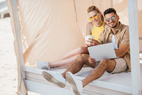 Feliz pareja joven en gafas de sol utilizando el ordenador portátil en bungalow - foto de stock