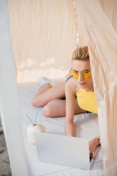 Hermosa mujer joven en gafas de sol utilizando el ordenador portátil mientras está acostado en bungalow - foto de stock