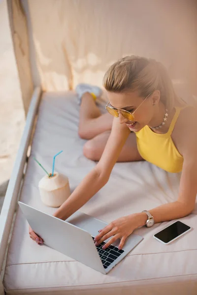 Vue grand angle de sourire belle fille dans les lunettes de soleil en utilisant un ordinateur portable tout en se reposant dans le bungalow — Photo de stock