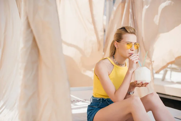 Beautiful young woman in sunglasses drinking refreshing cocktail while sitting in bungalow — Stock Photo