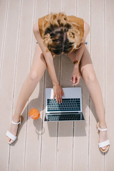 Vista aérea de la chica bonita usando el ordenador portátil mientras está sentado en la superficie de madera en el día soleado - foto de stock