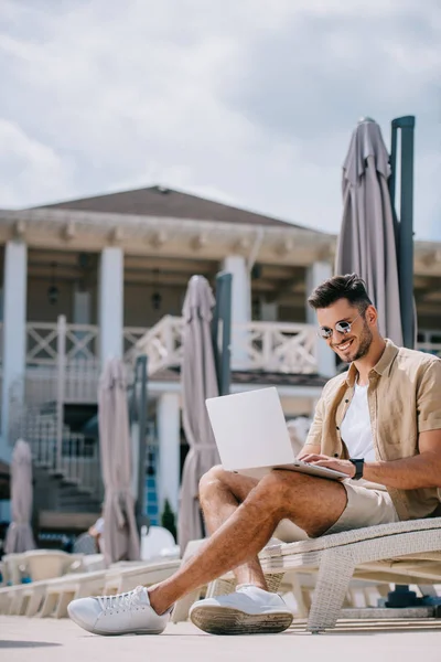 Улыбающийся молодой человек в солнечных очках, сидящий на шезлонге и использующий ноутбук — стоковое фото