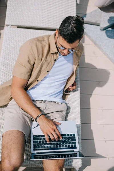 Hochwinkelaufnahme eines jungen Mannes mit Sonnenbrille, der auf einer Chaiselongue liegt und Laptop benutzt — Stockfoto