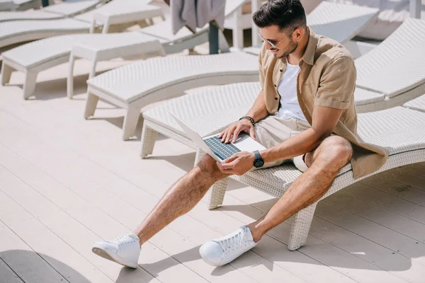 Bello giovane seduto sulla chaise longue e utilizzando il computer portatile a bordo piscina — Foto stock