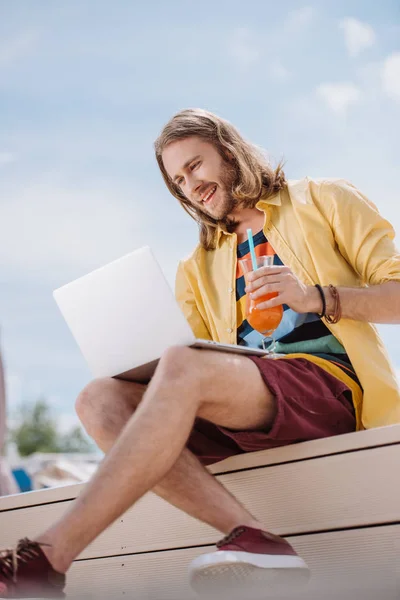 Joven sonriente sosteniendo cóctel y usando el ordenador portátil en la playa - foto de stock