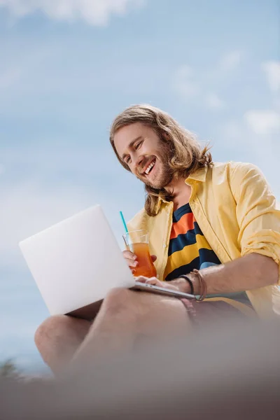 Vista de ángulo bajo del joven sonriente sosteniendo cóctel y usando el ordenador portátil en la playa - foto de stock