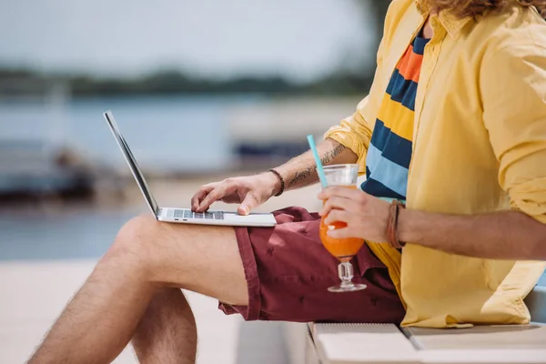 Cropped shot of young man holding cocktail and using laptop outdoors — Stock Photo