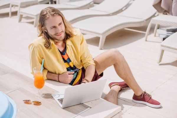 Високий кут зору усміхненого молодого чоловіка, який використовує ноутбук, відпочиваючи з коктейлем біля басейну — стокове фото