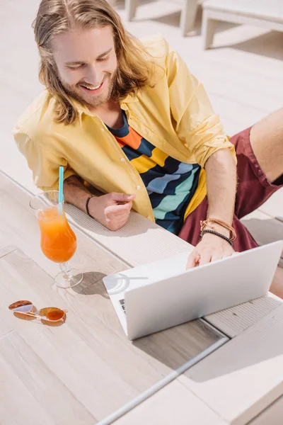 Joven sonriente usando el ordenador portátil mientras descansa con un cóctel en la piscina - foto de stock
