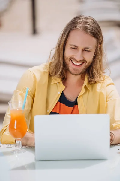 Joven sonriente usando el ordenador portátil mientras está sentado al aire libre en el día soleado - foto de stock