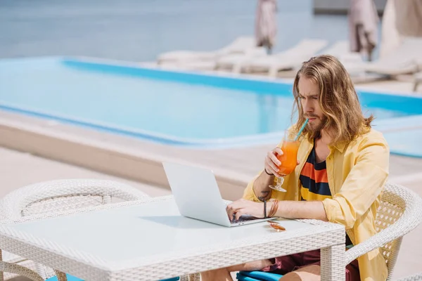 Jovem bonito usando laptop e beber coquetel perto da piscina — Fotografia de Stock