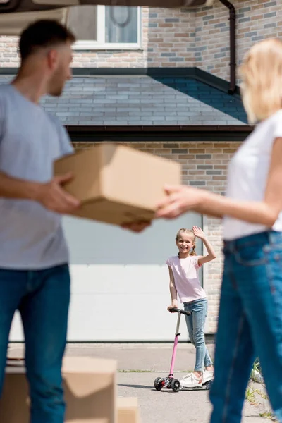 Hombre dando caja de cartón a la esposa mientras que su hija montar en patada scooter detrás en el patio de la nueva casa - foto de stock