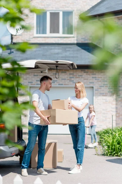 Homme donnant des boîtes en carton à la femme tandis que leur fille chevauchant sur trottinette derrière dans la cour de la nouvelle maison — Photo de stock