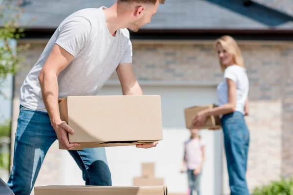 Close up view of man holding cardboard boxes while his wife and daughter standing behind in yard of new house — Stock Photo