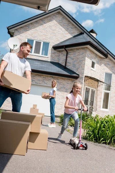 Smiling little kid riding on kick scooter and her parents unpacking cardboard boxes for relocation into new house — Stock Photo