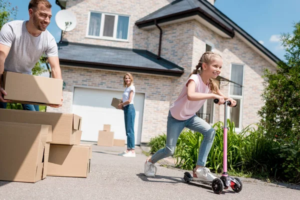 Little kid riding on kick scooter and her parents unpacking cardboard boxes for relocation into new house — Stock Photo
