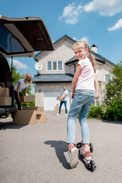 Criança feliz montando no chute de scooter e seus pais desembalando caixas de papelão para realocação em nova casa — Fotografia de Stock
