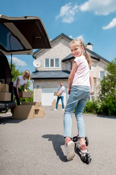 Niña feliz montando en patada scooter y sus padres desempacando cajas de cartón para la reubicación en la nueva casa - foto de stock