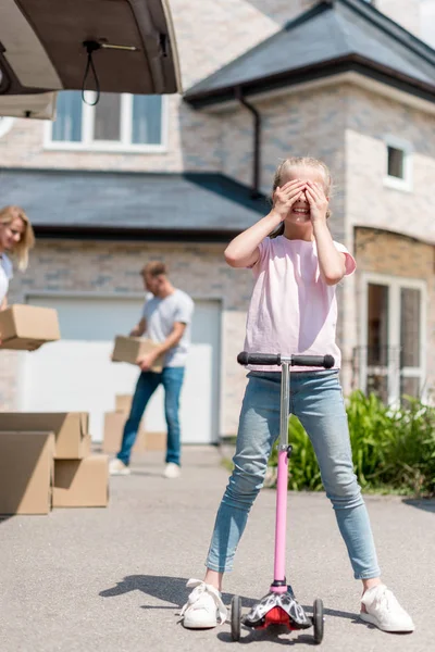 Little kid covering eyes on kick scooter and her parents unpacking cardboard boxes for relocation into new house — Stock Photo