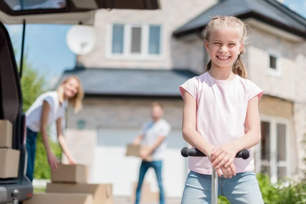 Smiling little kid on kick scooter and her parents unpacking cardboard boxes for relocation into new house — Stock Photo