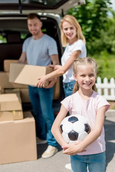 Happy little kid holding soccer ball while her parents unpacking cardboard boxes for relocation — Stock Photo