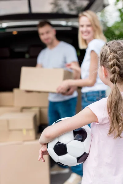 Plan recadré d'un petit enfant tenant un ballon de football pendant que ses parents déballent des boîtes en carton pour le déménagement — Photo de stock